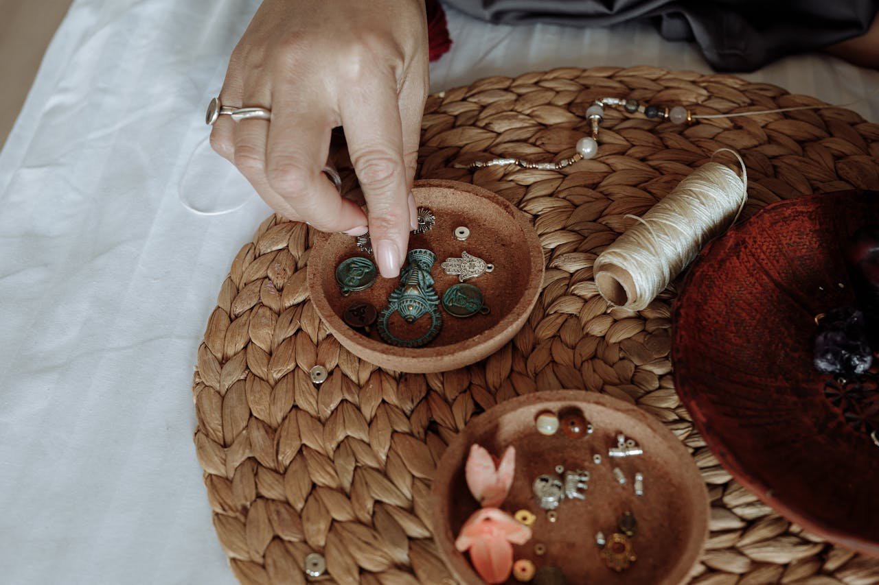 Close-up of a hand organizing jewelry components on a woven mat with thread and wooden bowls.